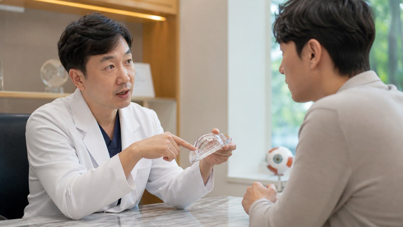 밝은 진료실에서 의료진이 투명한 각막 모형을 들고 내원자에게 스마일라식 방식을 설명하는 모습